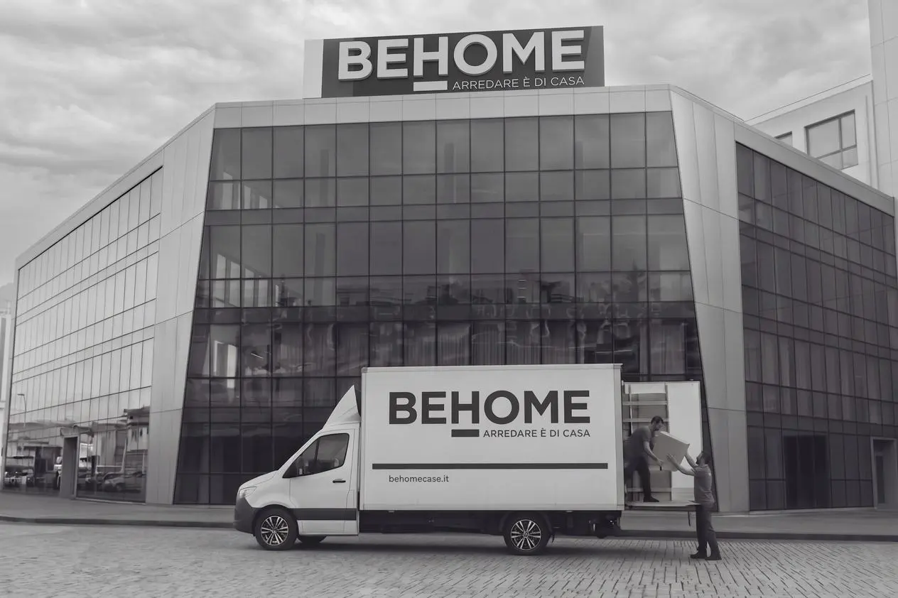 Camion per le consegne con logo BEHOME parcheggiato davanti a uno showroom del brand, con due operatori logistici al lavoro, a dimostrazione dell'efficiente servizio di trasporto e montaggio garantito in tutta Italia ed Europa.